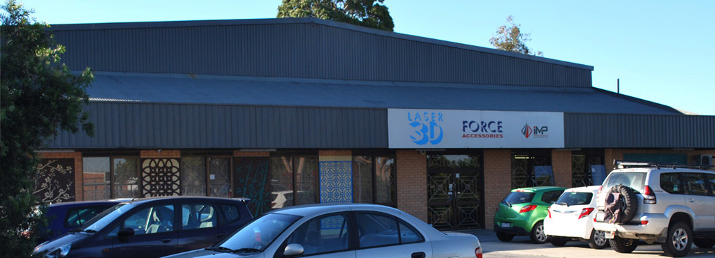 A building with a sign with 3 logos: Laser 3D, Force accessories, and iMP. There are cars parked outside the building in a carpark.