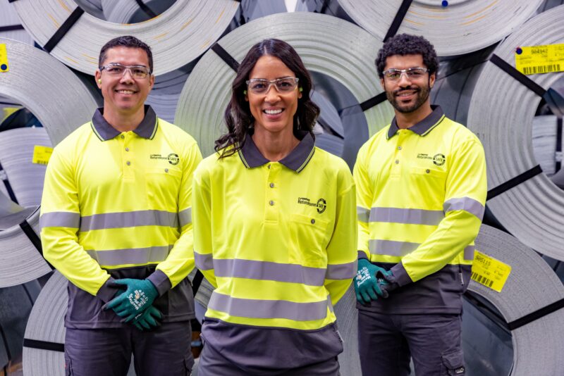 Laser 3D team members in high-visibility PPE standing in front of stacked metal coils.