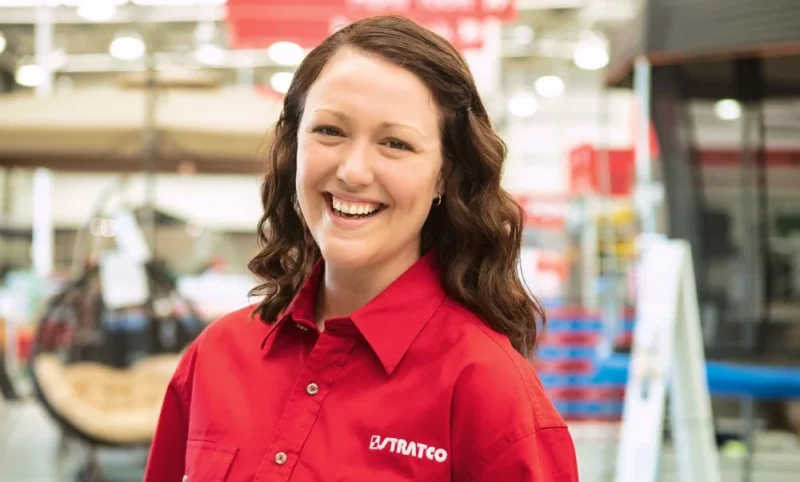 Smiling female employee wearing a red Stratco work shirt standing inside an industrial facility.”