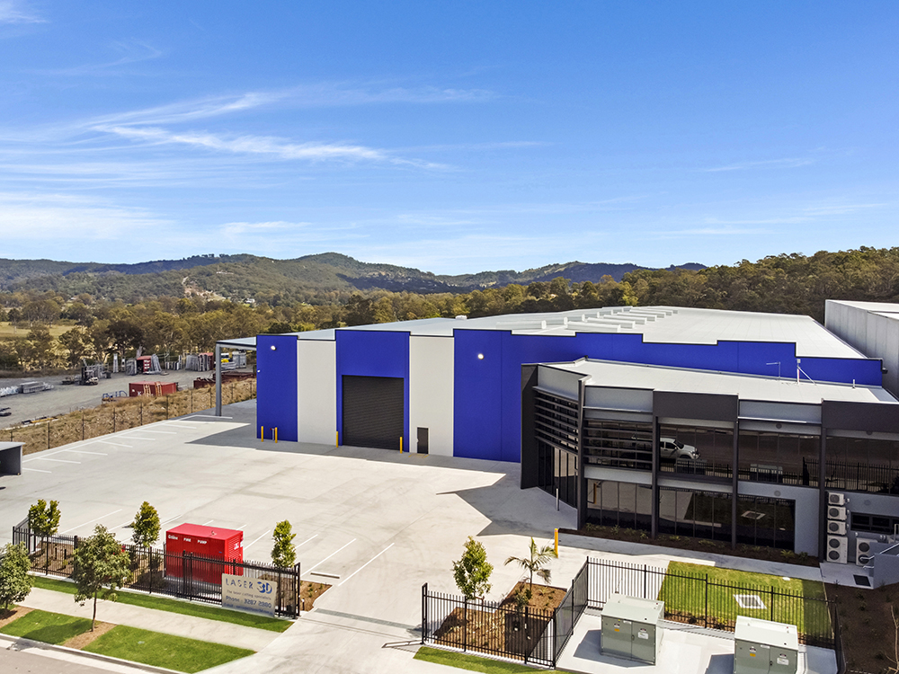 A very large warehouse with attached office space and large carpark as seen from above.
